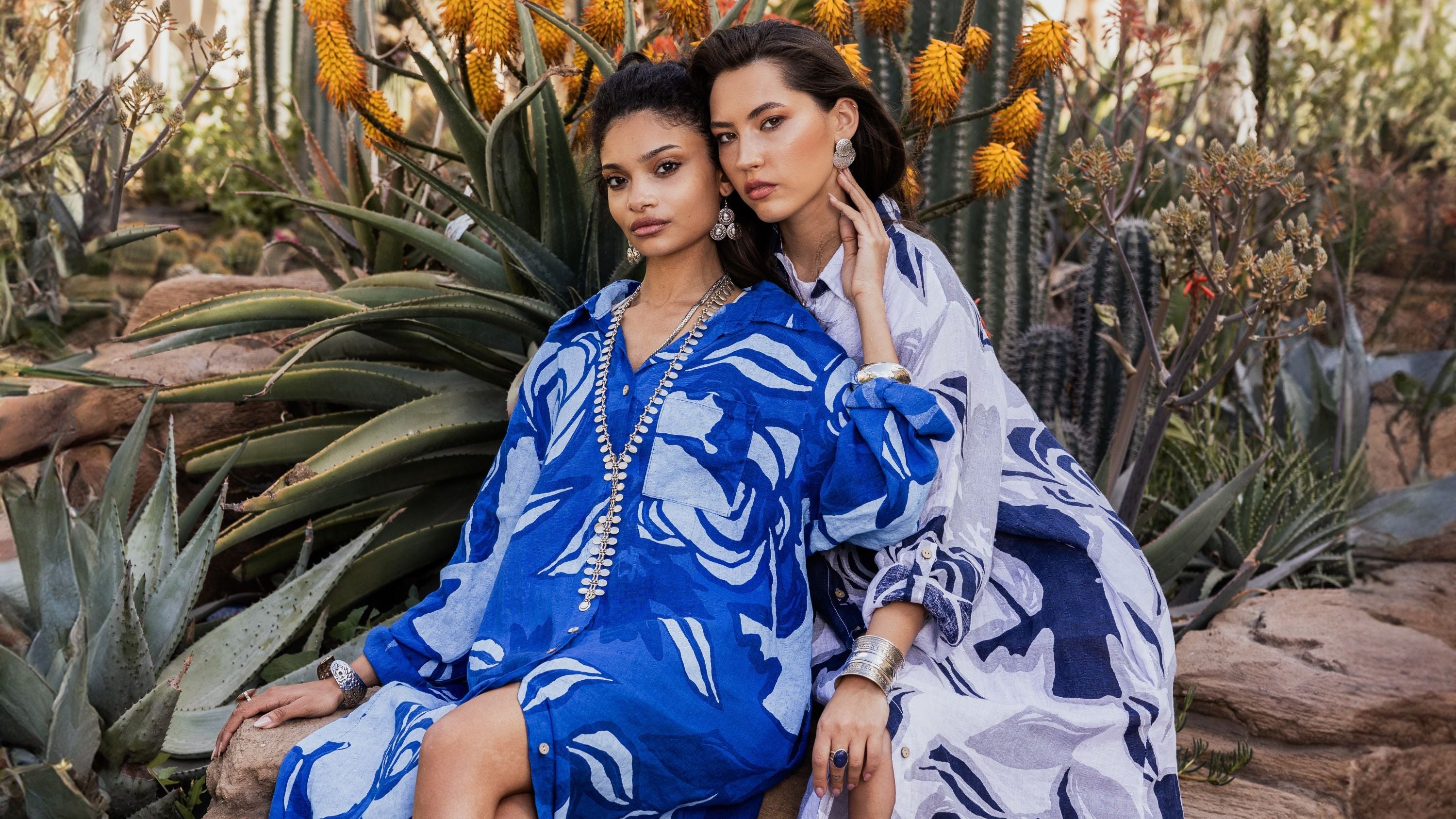 Two women in blue and white patterned dresses sitting among cacti.